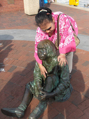 4 photos show life-sized statues on a plaza, one is of Haley sitting on a wall reading a book, there are 3 other statures of children listening to him.  One child statue lies on his stomach looking up at Haley, two others are sitting down.  Standing on Haley's lap and on one of the child statues is a real child.